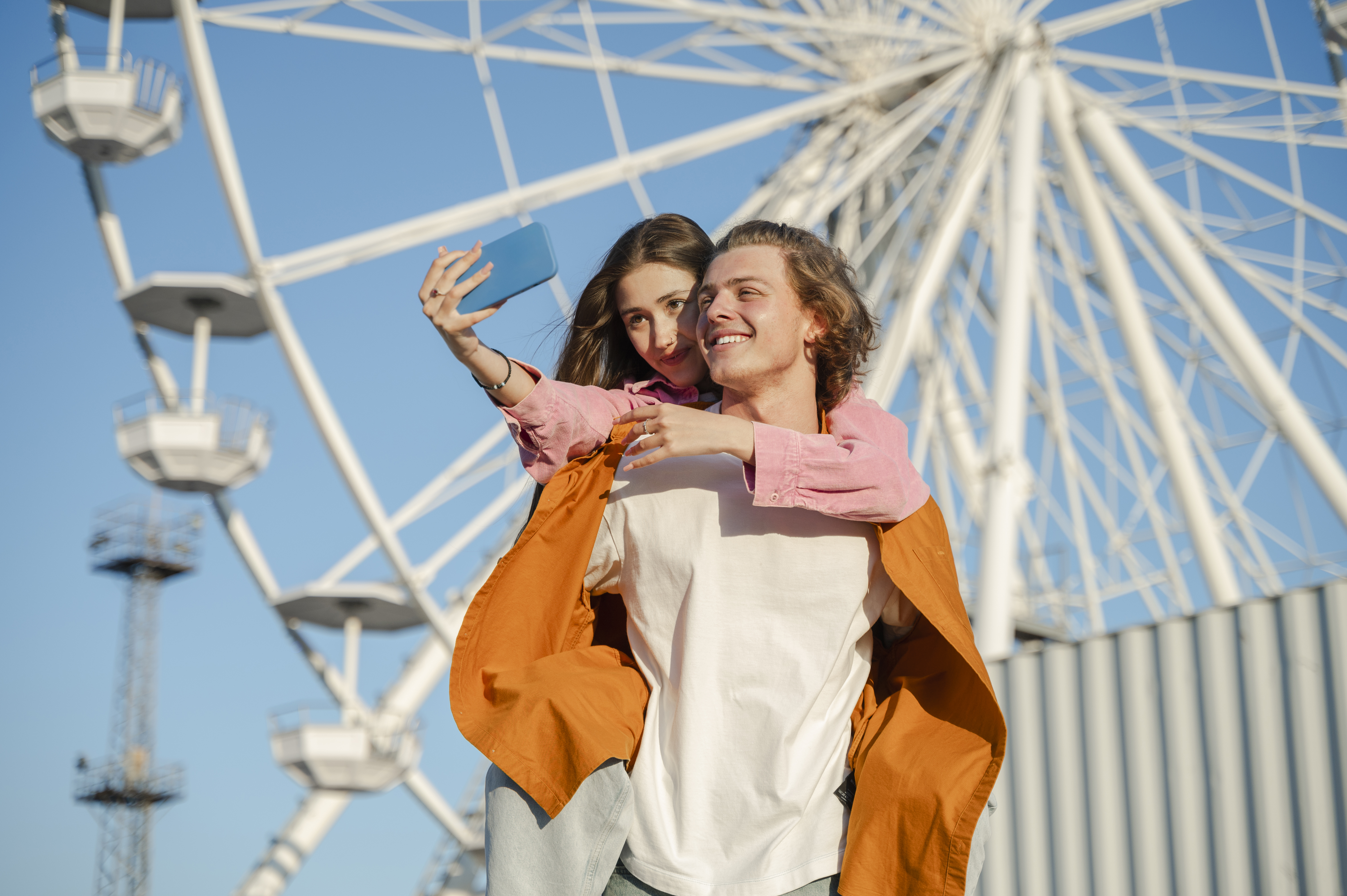 Smiling couple clicking selfie in front of ferris wheel at amusement park